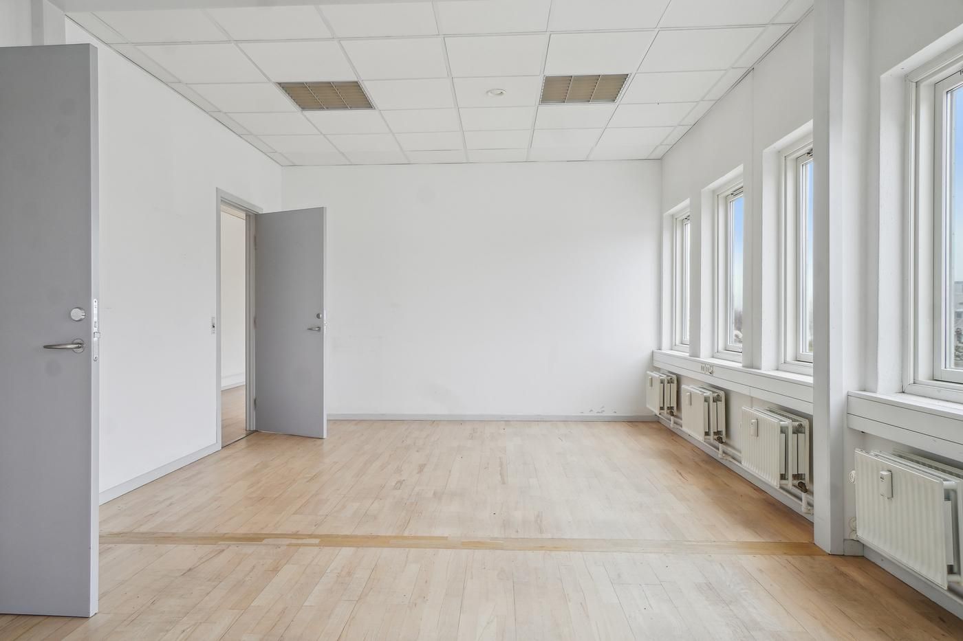 En tom kontorbygning med hvide vægge, hårdt trægulv og hvide lofter med store ventiler. Det er åbne døre og vinduer i rummet.