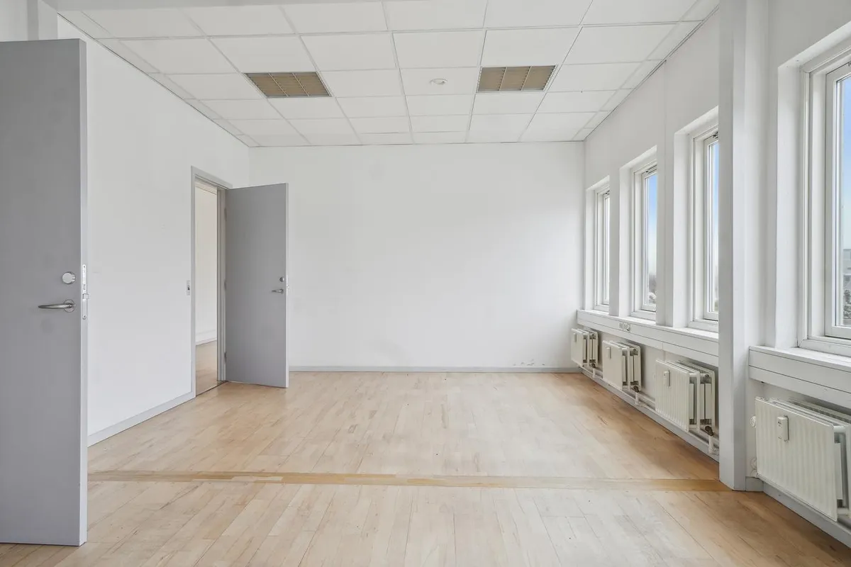En tom kontorbygning med hvide vægge, hårdt trægulv og hvide lofter med store ventiler. Det er åbne døre og vinduer i rummet.
