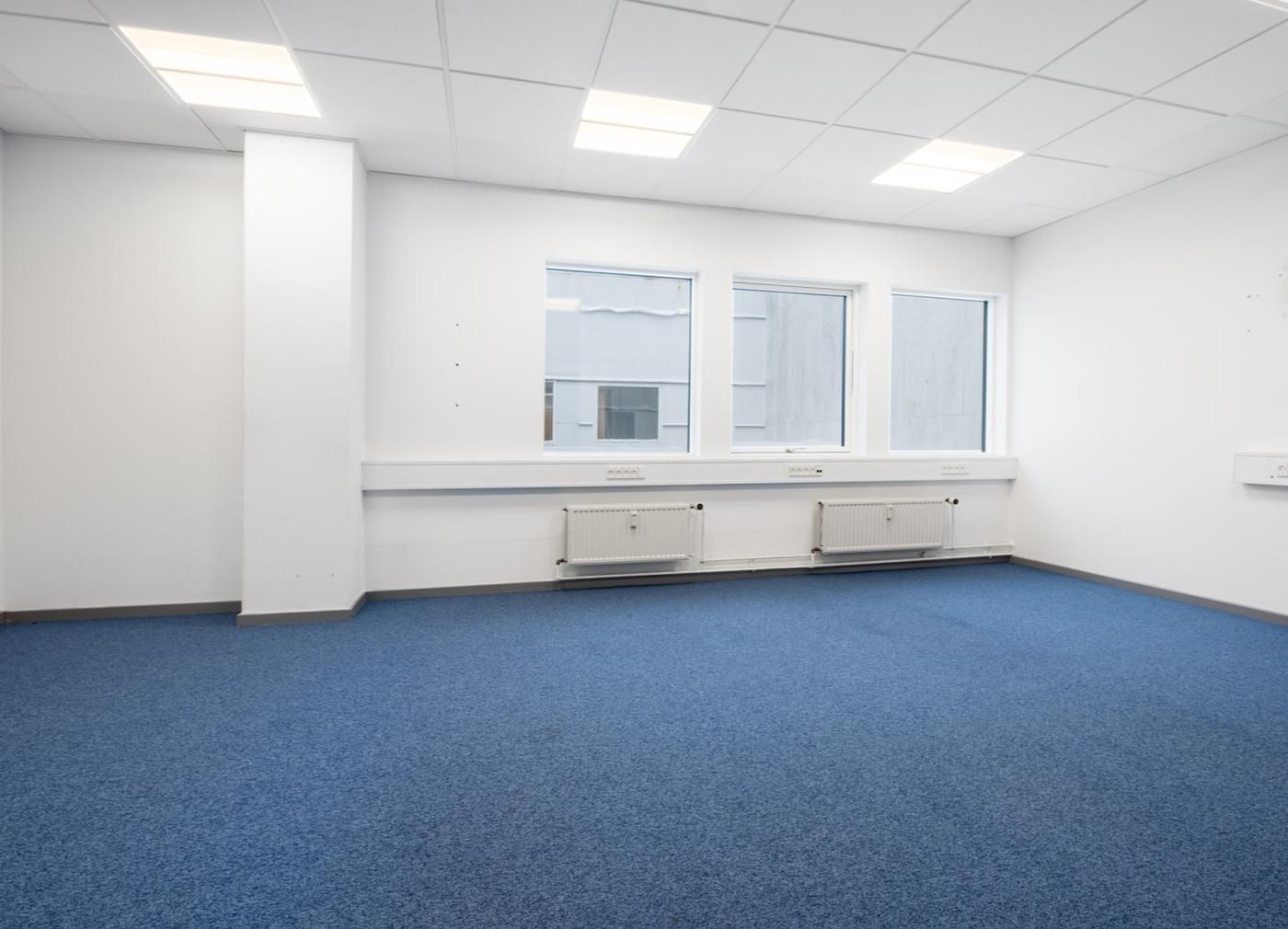 En stor, lyse og rummelig kontor med lyseblå gulv og hvide vægge, fire vinduer og tre radiatorer i en række på væggen