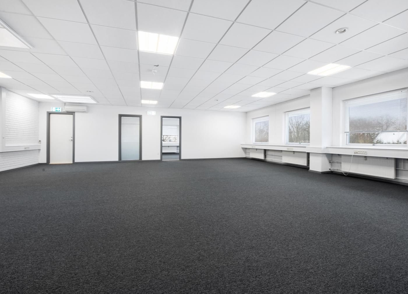 Måske en kontorlokal med hvide vægge og gulv, lyse hvide lofter, fire døre og fire vinduer med jalousier, hvor man ser træer