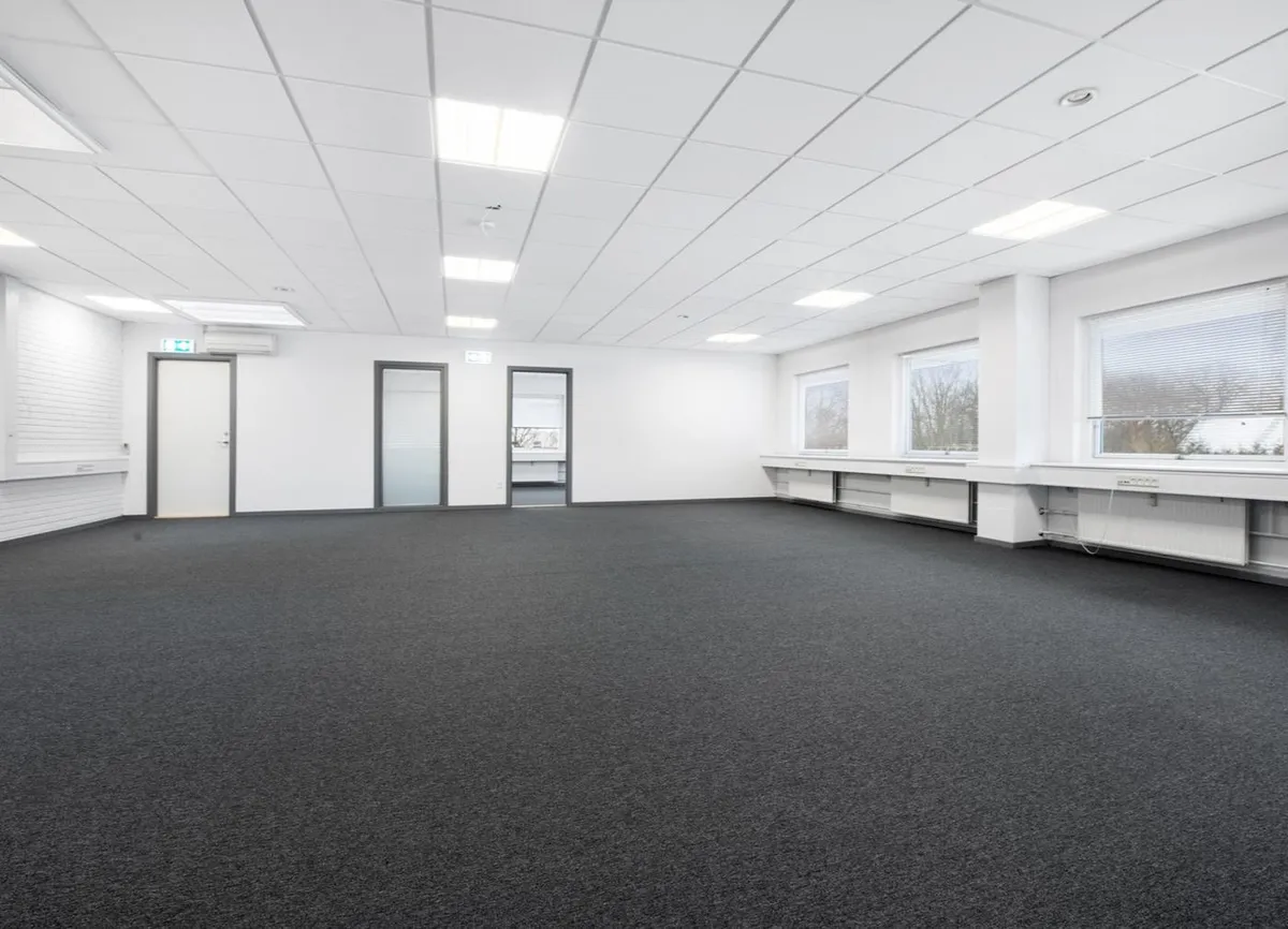 Måske en kontorlokal med hvide vægge og gulv, lyse hvide lofter, fire døre og fire vinduer med jalousier, hvor man ser træer