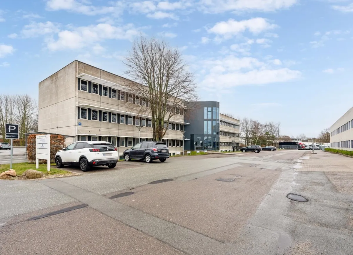 Tre parkerede biler og en vejskilt i en parkeringsplads foran et stort kontorbyggeri med glasvinduer og et rektangulært