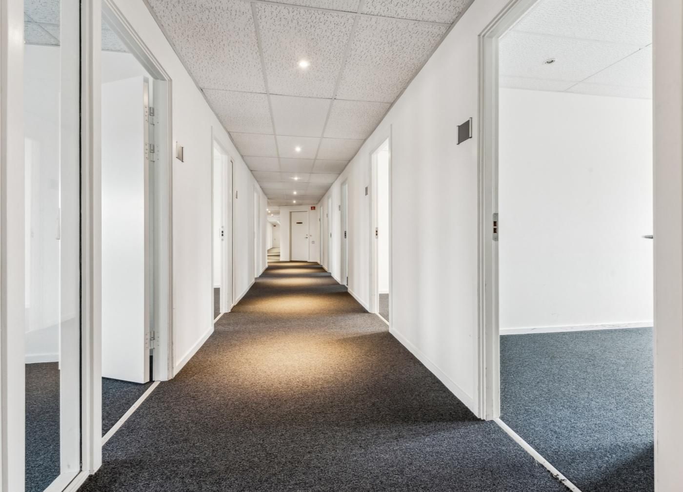 En langstrakt og smal gang med lyse hvide vægge og lysegrå gulvbelægning er oplyst med hvide loftslamper
