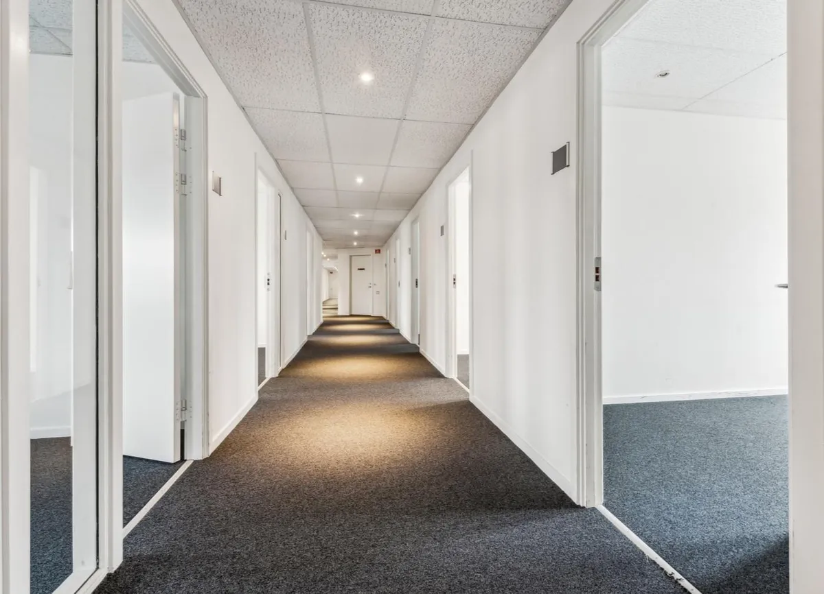 En langstrakt og smal gang med lyse hvide vægge og lysegrå gulvbelægning er oplyst med hvide loftslamper