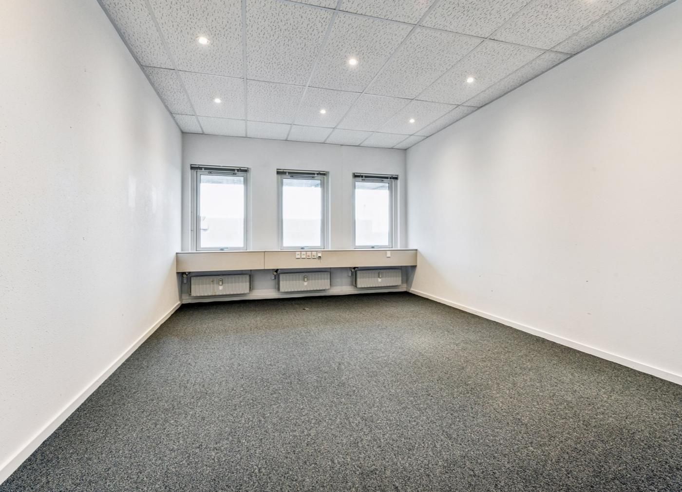 En tom og veludstyret kontorrum med tre vinduer, lyse og hvide vægge, og gulvbelægning i gråt tekstil