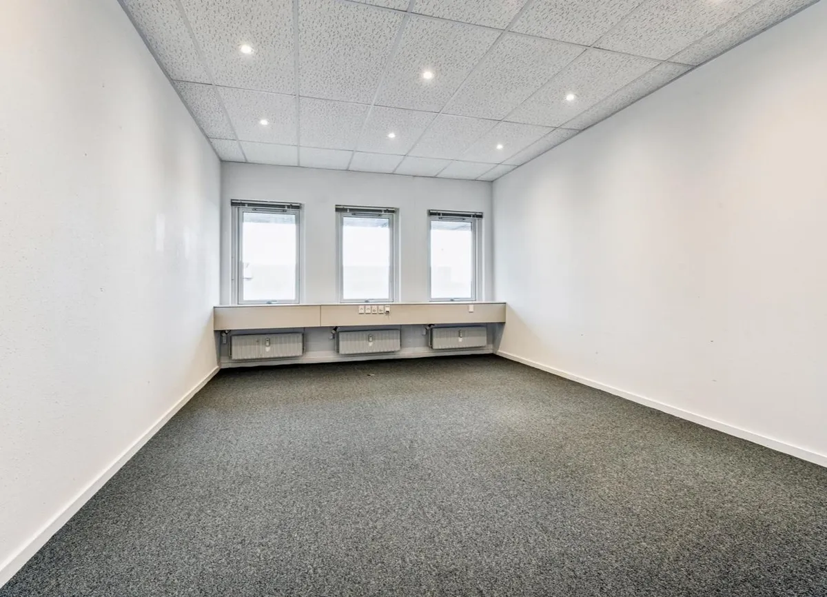 En tom og veludstyret kontorrum med tre vinduer, lyse og hvide vægge, og gulvbelægning i gråt tekstil