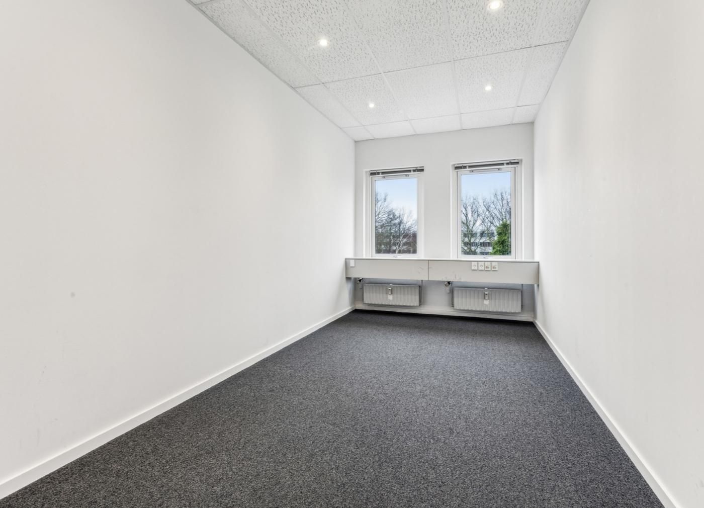 Et tomt kontorrum med hvide vægge, lyseblå gulvbelægning og to vinduer med udsigt til grønne træer