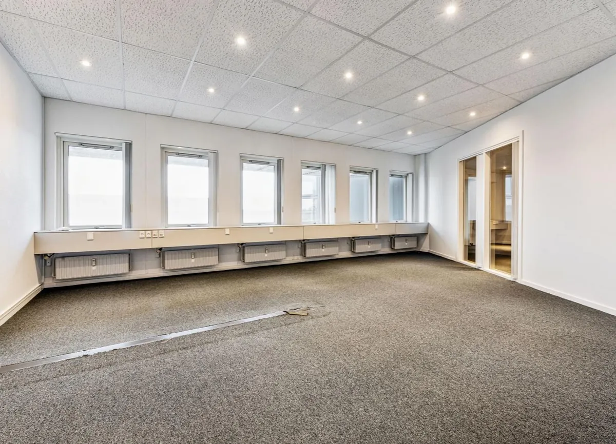 Stort, lyst kontorlokal med hvide vægge og gulvbelægning samt fire store vinduer på en side