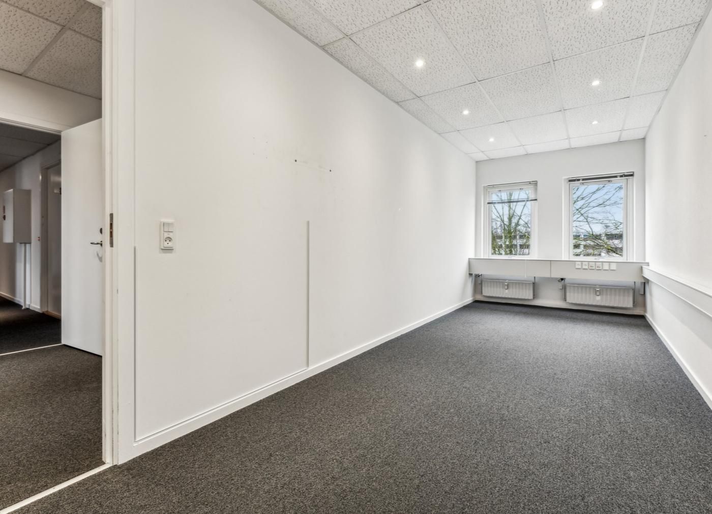 Lyst og rummeligt kontorlokal med tavle, vinduer, radiatorer og hvide vægge og gulv