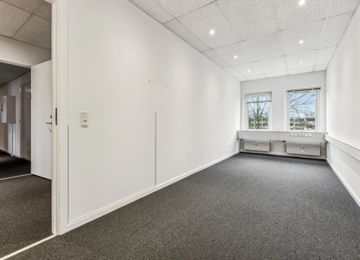 Lyst og rummeligt kontorlokal med tavle, vinduer, radiatorer og hvide vægge og gulv