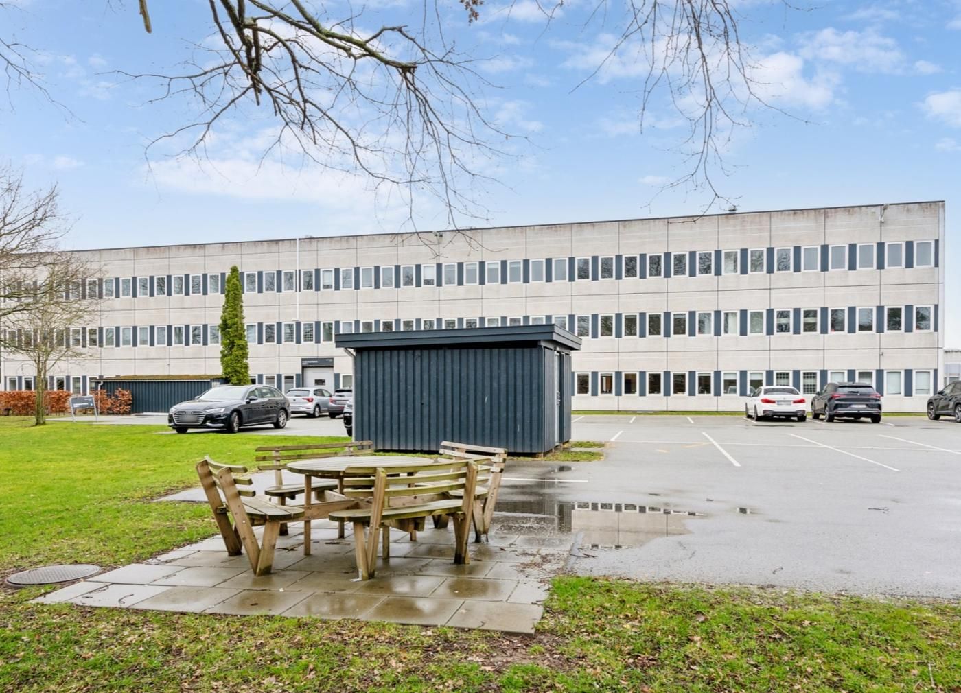 Træstole og bord udenfor en stor kontorbygning med parkeringsplads og biler i baggrunden