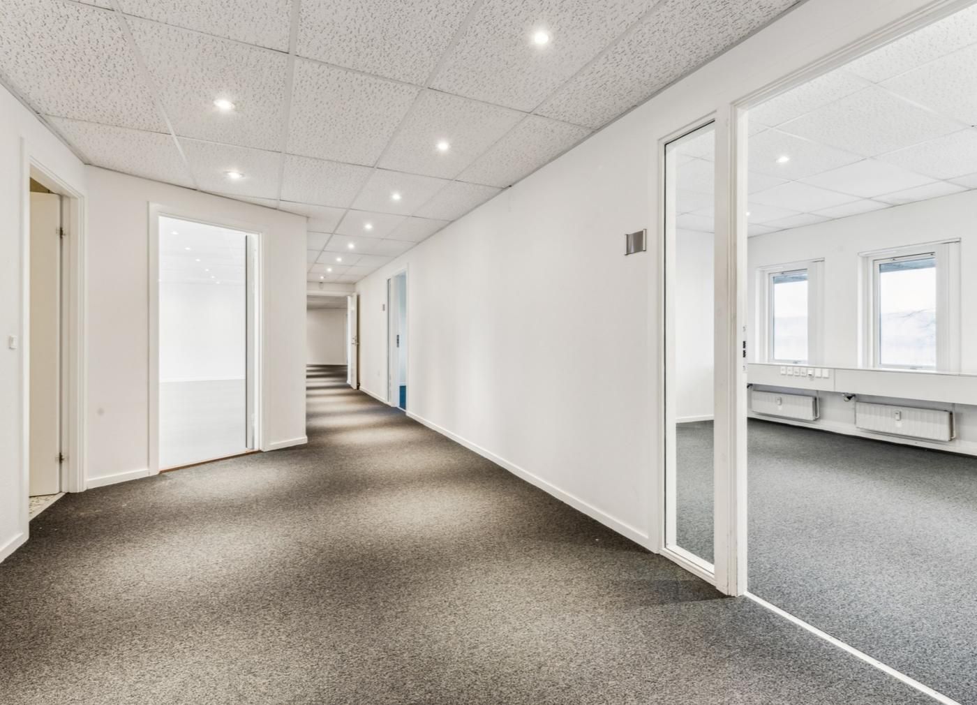 Kontorrum med hvide vægge, lysegul gulvbelægning og flere vinduer. Der er højt til loftet og flere døre i rummet.