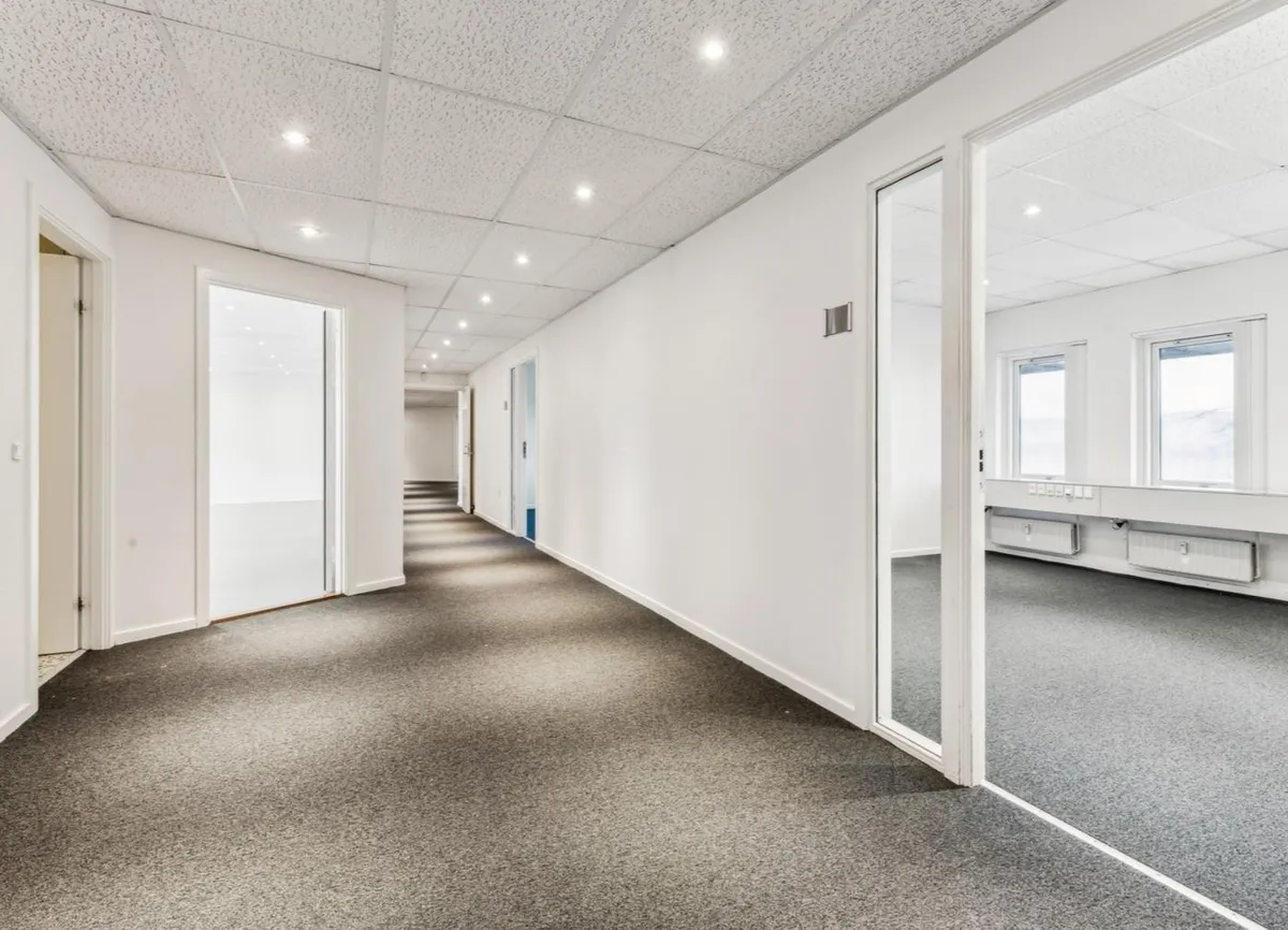 Kontorrum med hvide vægge, lysegul gulvbelægning og flere vinduer. Der er højt til loftet og flere døre i rummet.