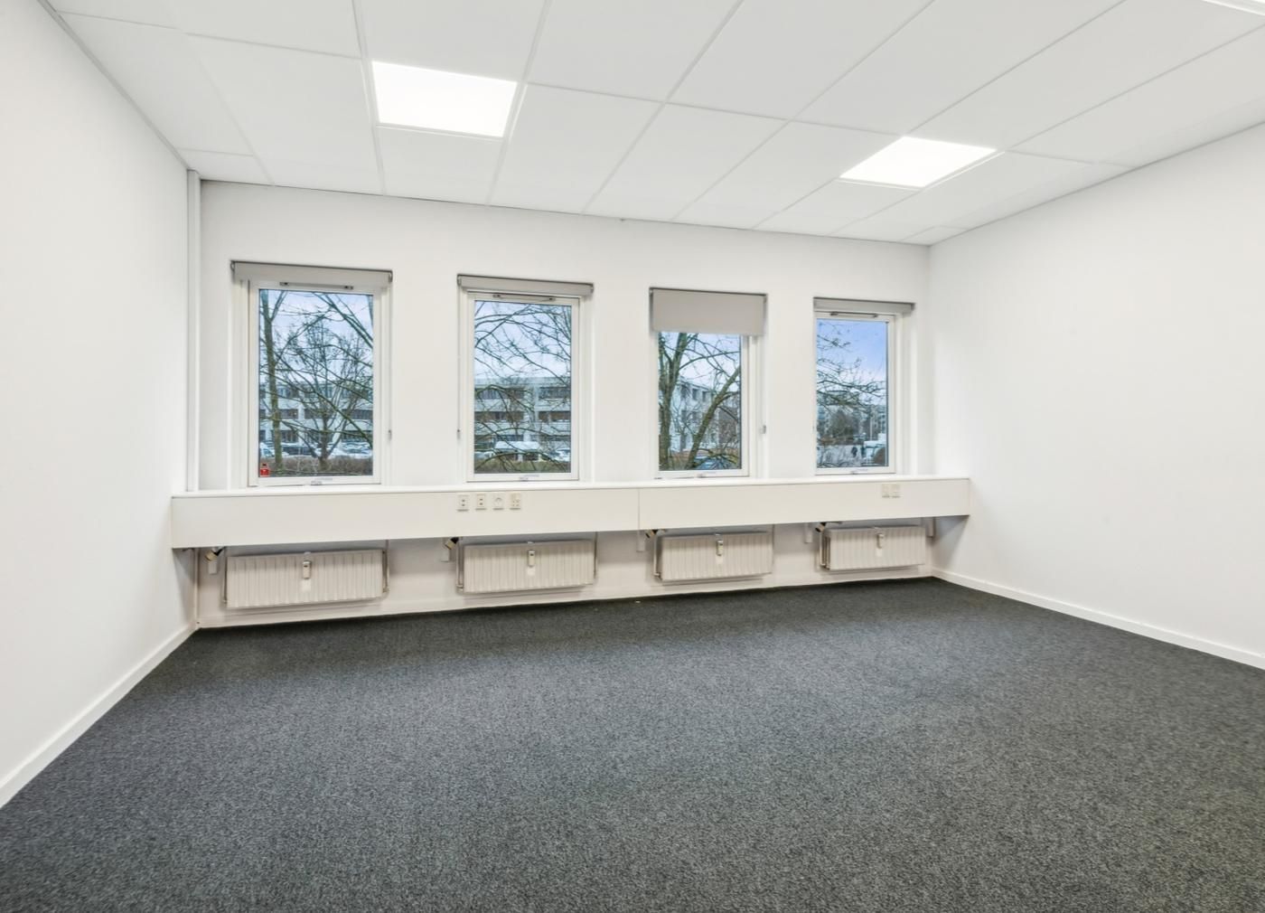 En tom kontor med tre vinduer og hvide vægge samt gulvbelægning i gråt. Der er radiatorer og vinduesgardiner.