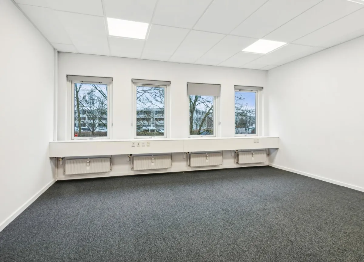 En tom kontor med tre vinduer og hvide vægge samt gulvbelægning i gråt. Der er radiatorer og vinduesgardiner.