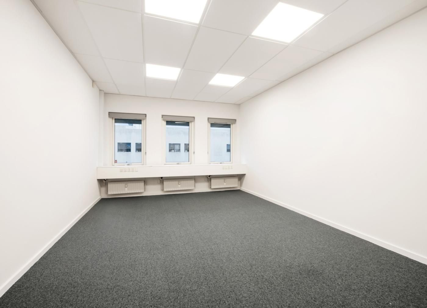 Rummelig, hvidt kontorlokal med lyse vinduer og lysekroner på loftet. Under vinduet er der et vindueskarm og tre radiatorer.