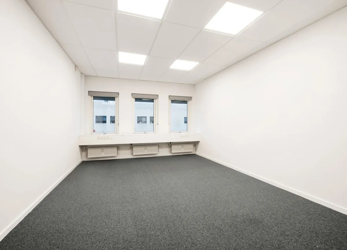 Rummelig, hvidt kontorlokal med lyse vinduer og lysekroner på loftet. Under vinduet er der et vindueskarm og tre radiatorer.