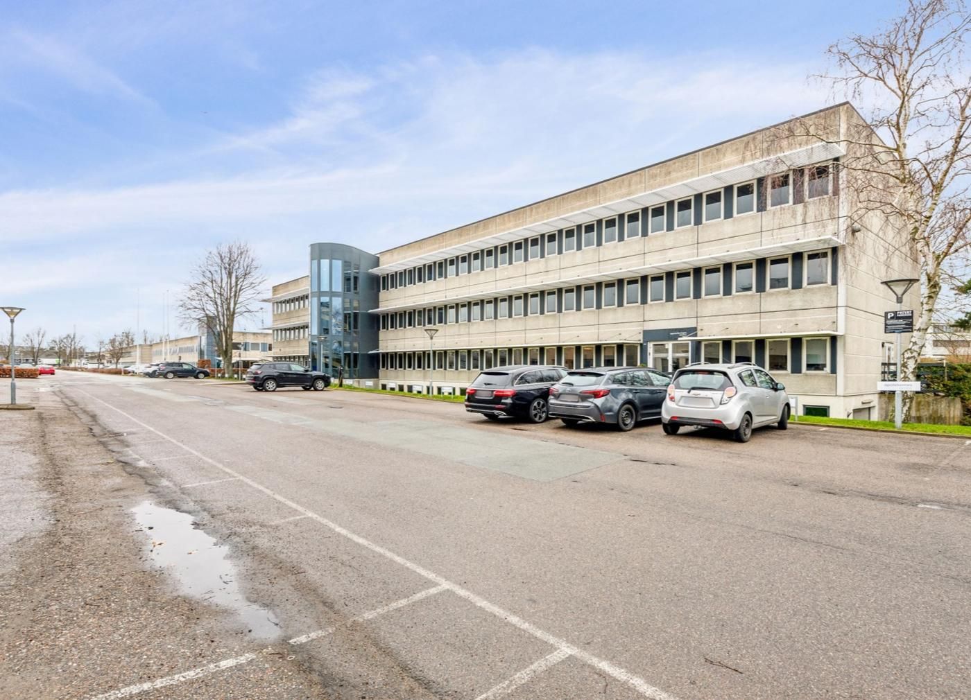 Kontorbyggeri med glasfacade og parkeringspladser samt biler, træer og grønne anlæg i baggrunden