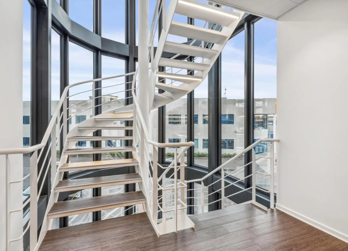 Et slankt designet trappe med hvidt stålgelændere og trætrin forbinder gulvet med et moderne kontorområde med store