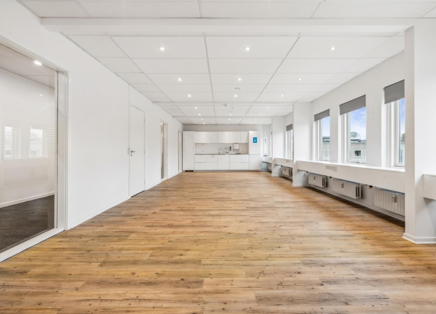 Lyse kontorlokaler med hvide vægge og lysegrå loft. Værelse med trægulv og store vinduer. Der er hvide køkkenskabe og