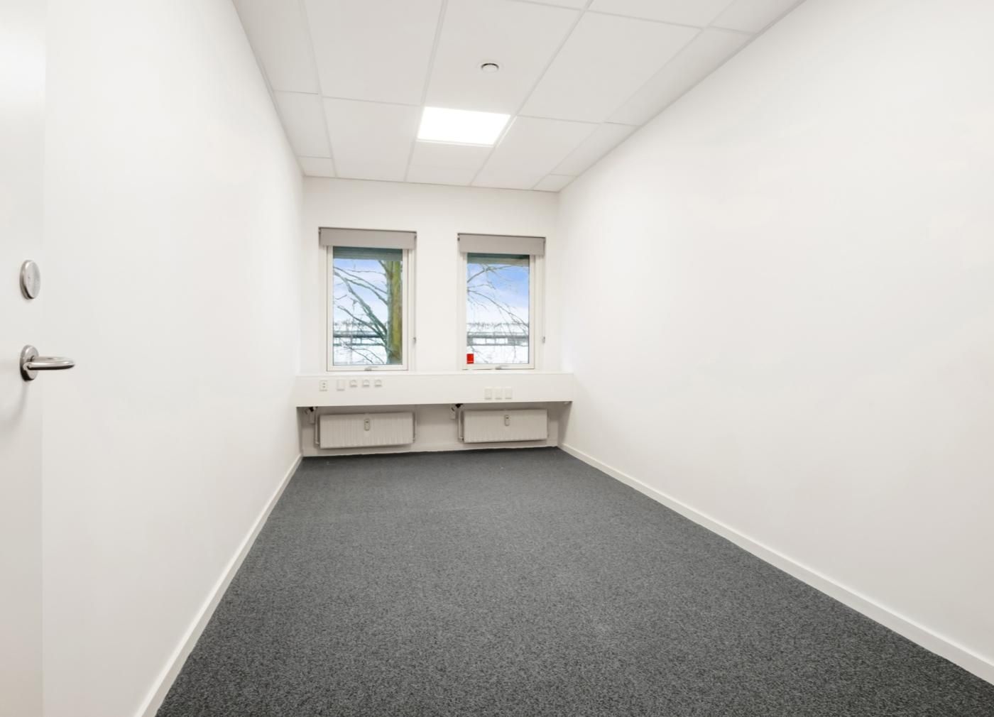 Moderne kontorlokale med hvide vægge og lysegule gulvbelægning, to store vinduer og radiatorer i væggen.