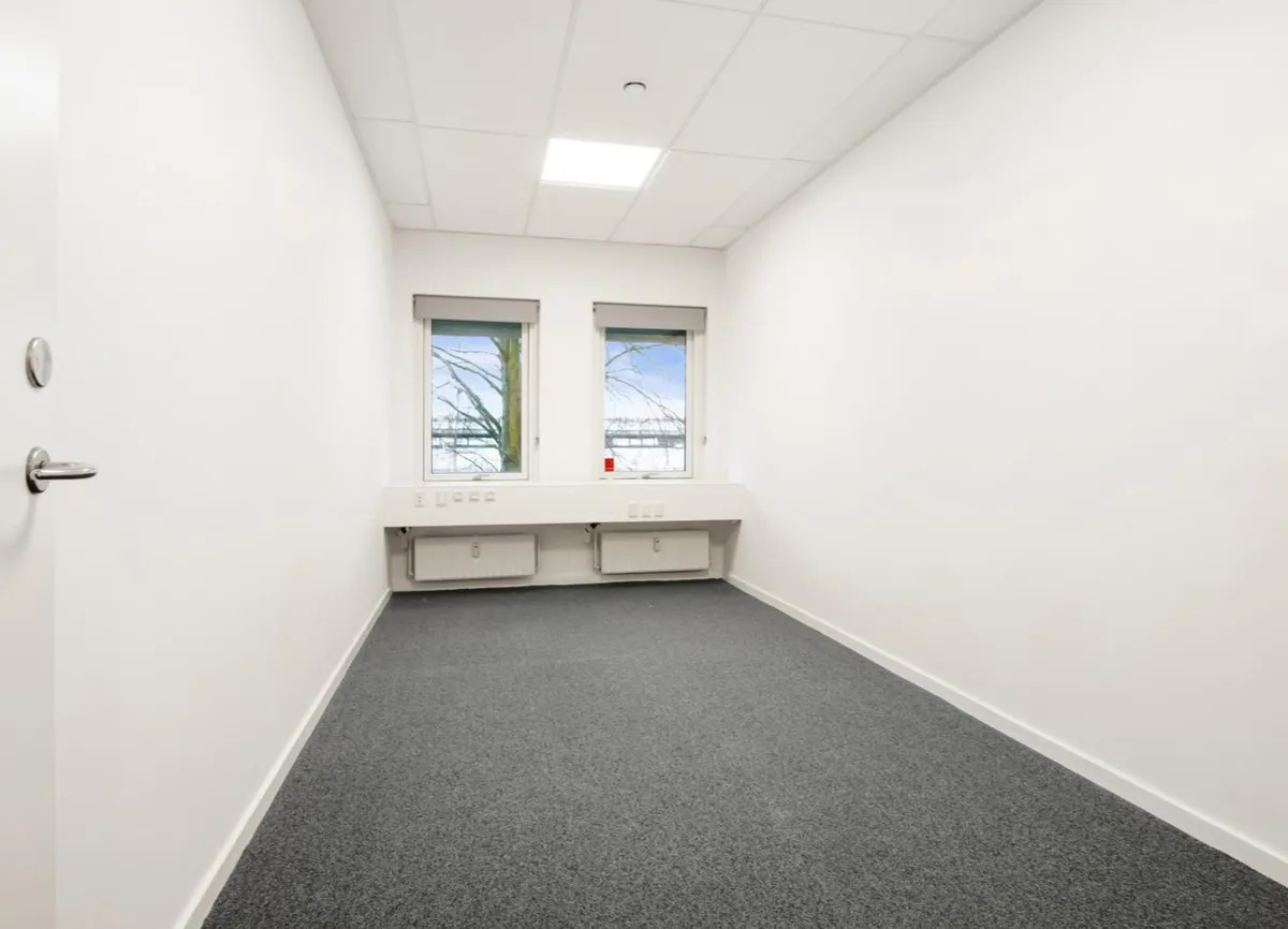 Moderne kontorlokale med hvide vægge og lysegule gulvbelægning, to store vinduer og radiatorer i væggen.
