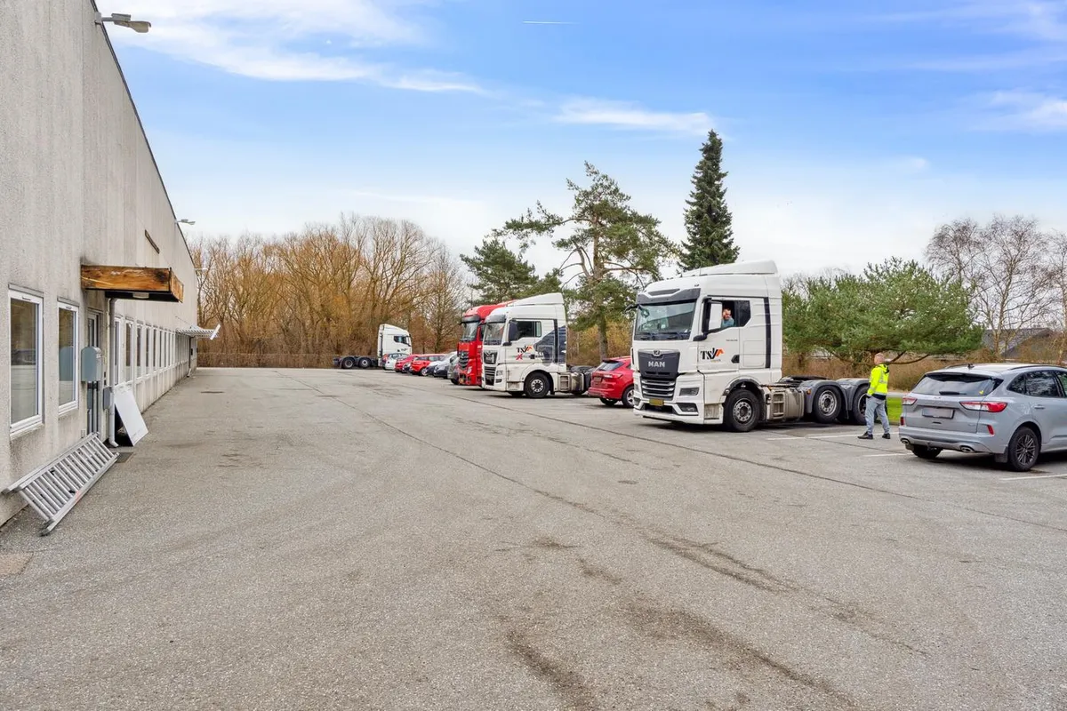 Lagerbygning med parkeringsplads, hvor flere lastbiler er parkeret, og en mand går mod en grå forhøjede bil