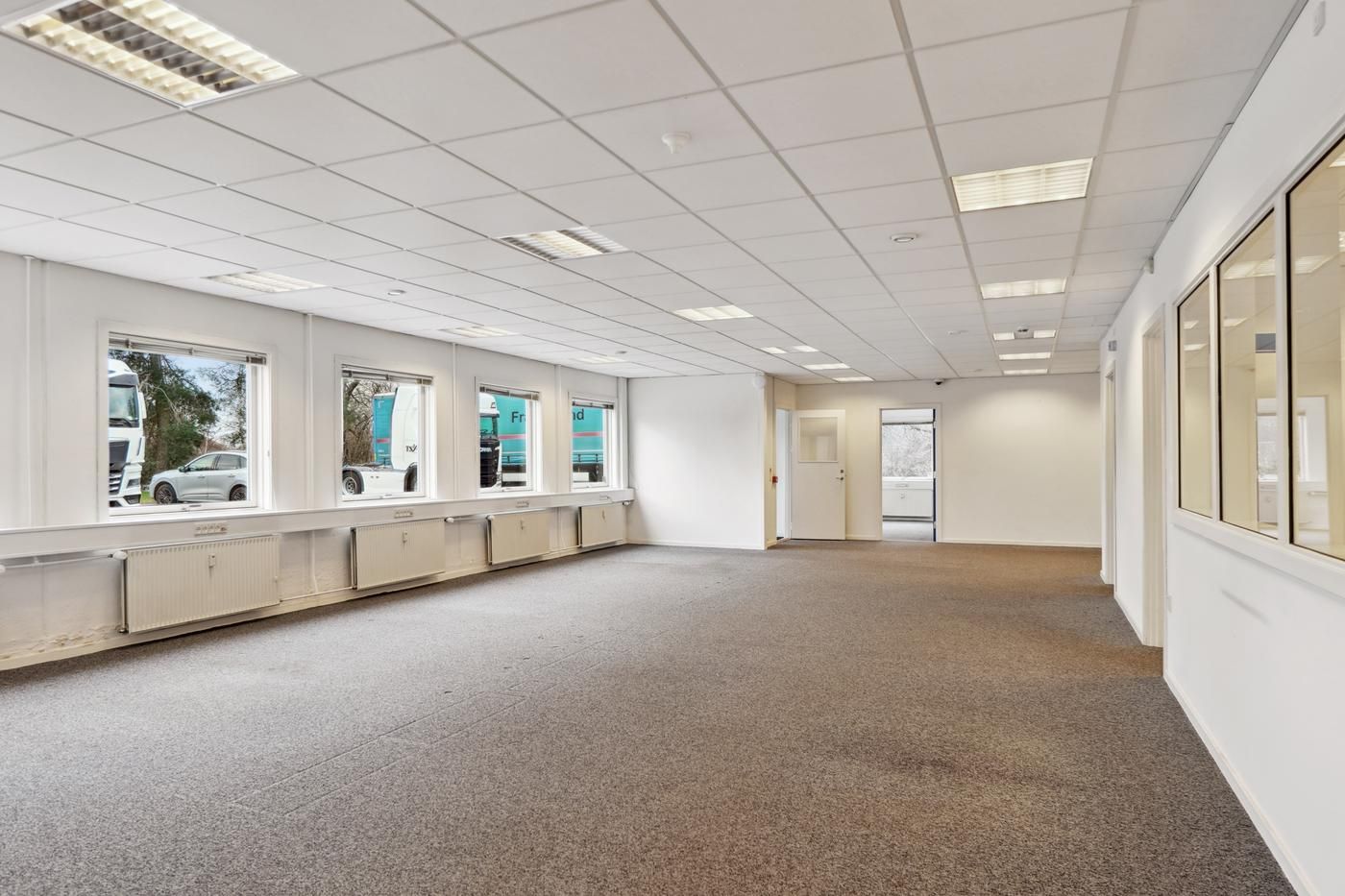 Store vinduer og hvide vægge i velholdt lokale med gardiner og møbler samt en åben dør og tekst på vinduet