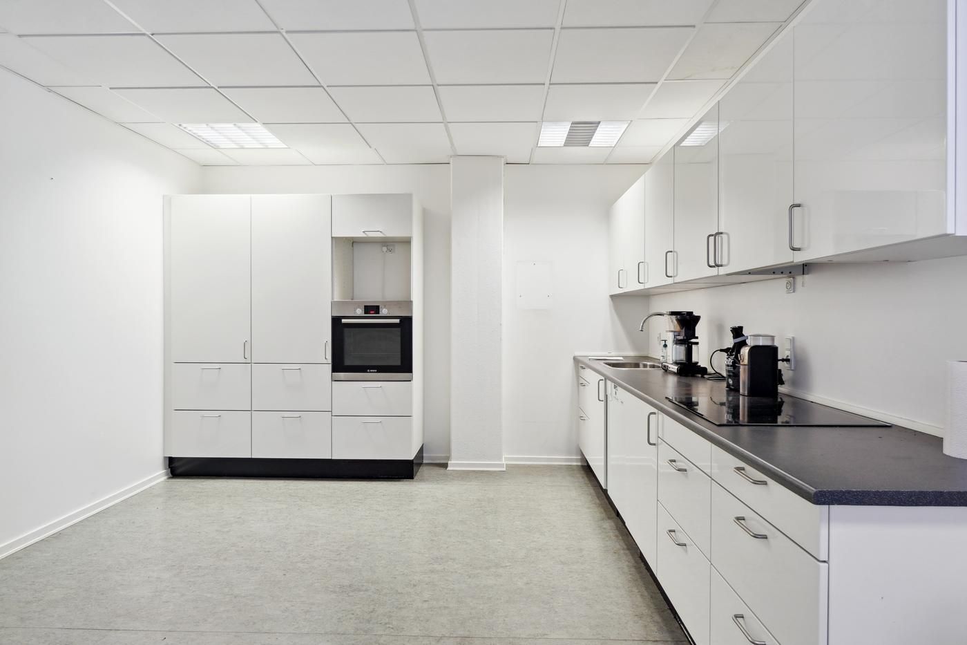 En tom kontorkøkken med hvide vægge, højloft og køkkenudstyret og -møbler i hvid og sort.