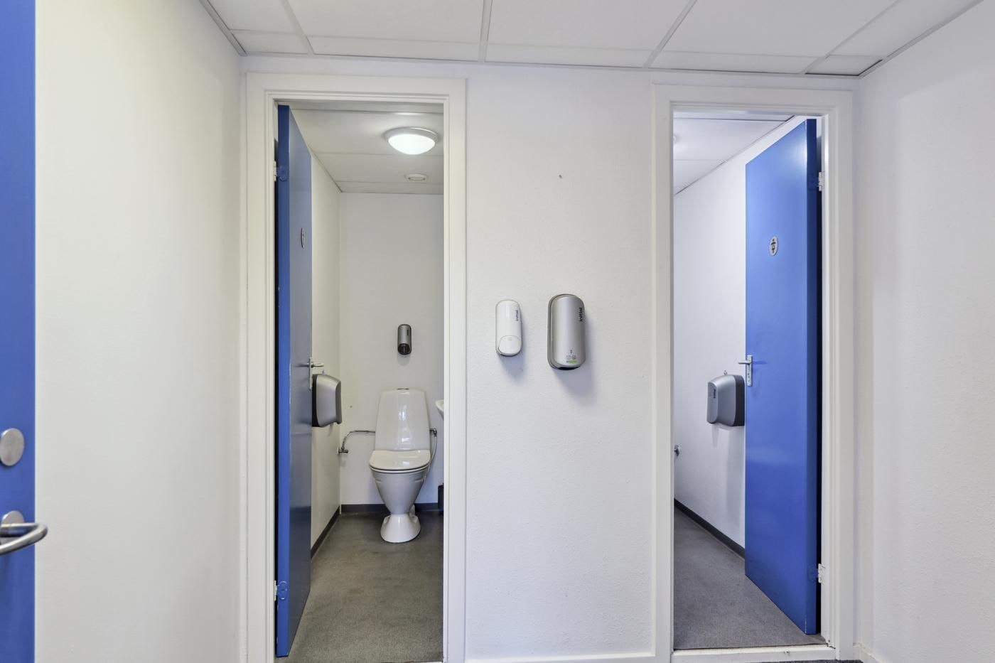 To toiletrum med hvide vægge og blå døre samt moderne toiletfaciliteter og håndvask med hånddesinfektionsmiddeluddeler