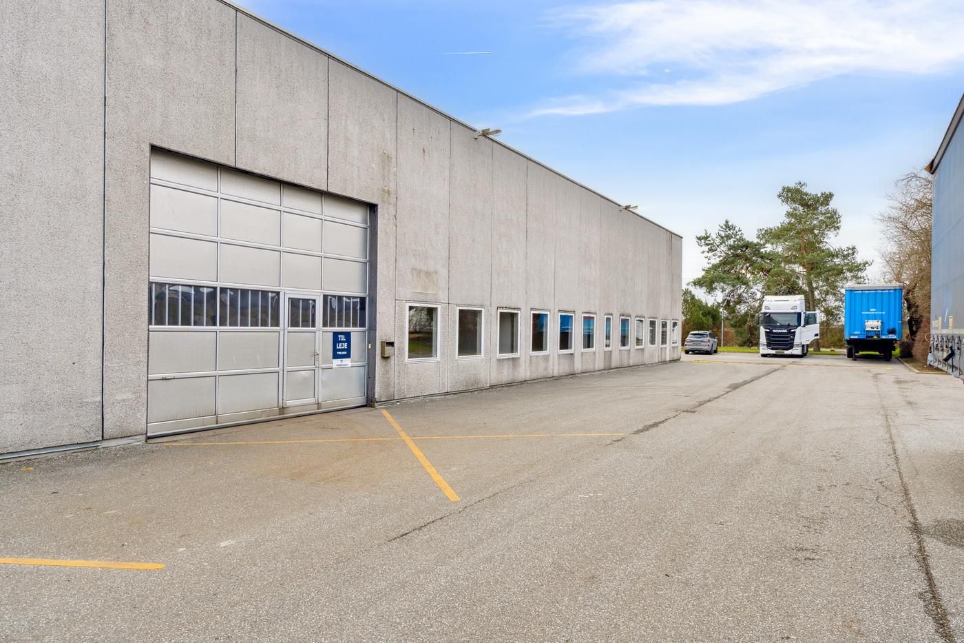 Lager med garagedør i lysegrå betonbygning, to lastbiler parkeret, to små biler og flere træer på siden