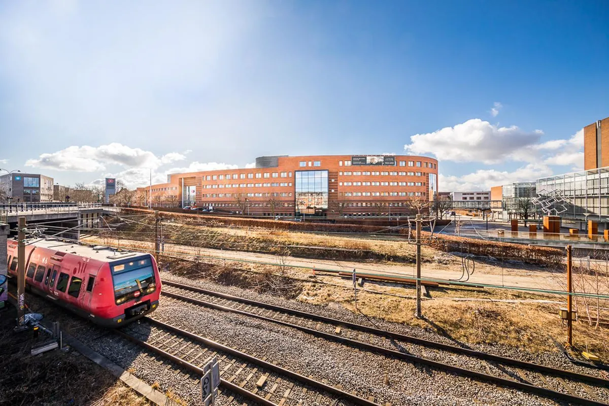 En rødt tog kører på skinnerne, og der er en stor bygning med flere vinduer bagved