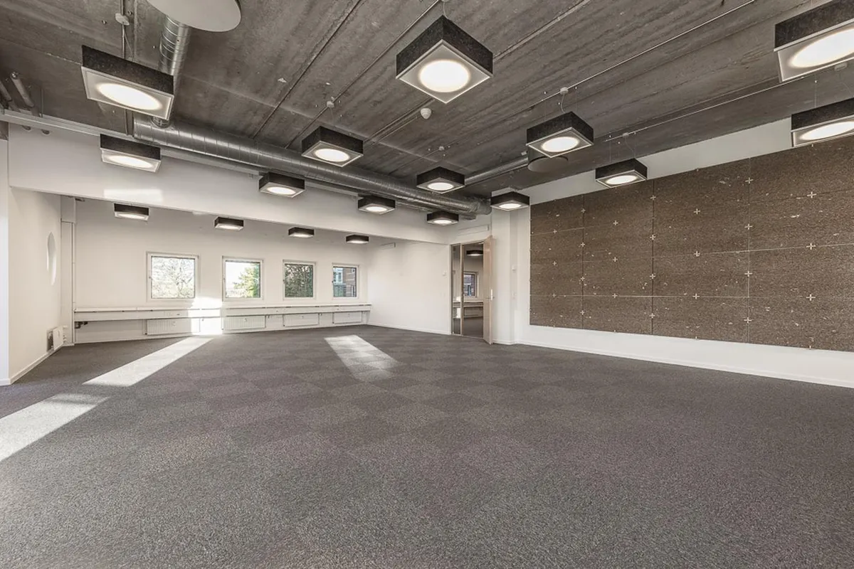 En stor og rummelig kontorlokal med lyse loftslamper, gulvtæppe og store ruder med udsigt til gade