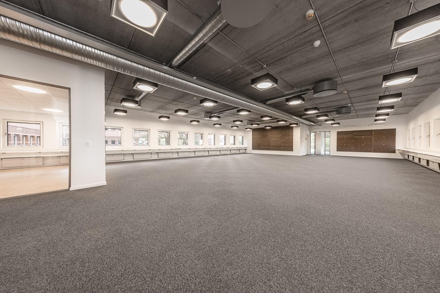 Stort kontor med karpetgulv og højt loft, hvor der er en stor åbning til et lille rum i baggrunden