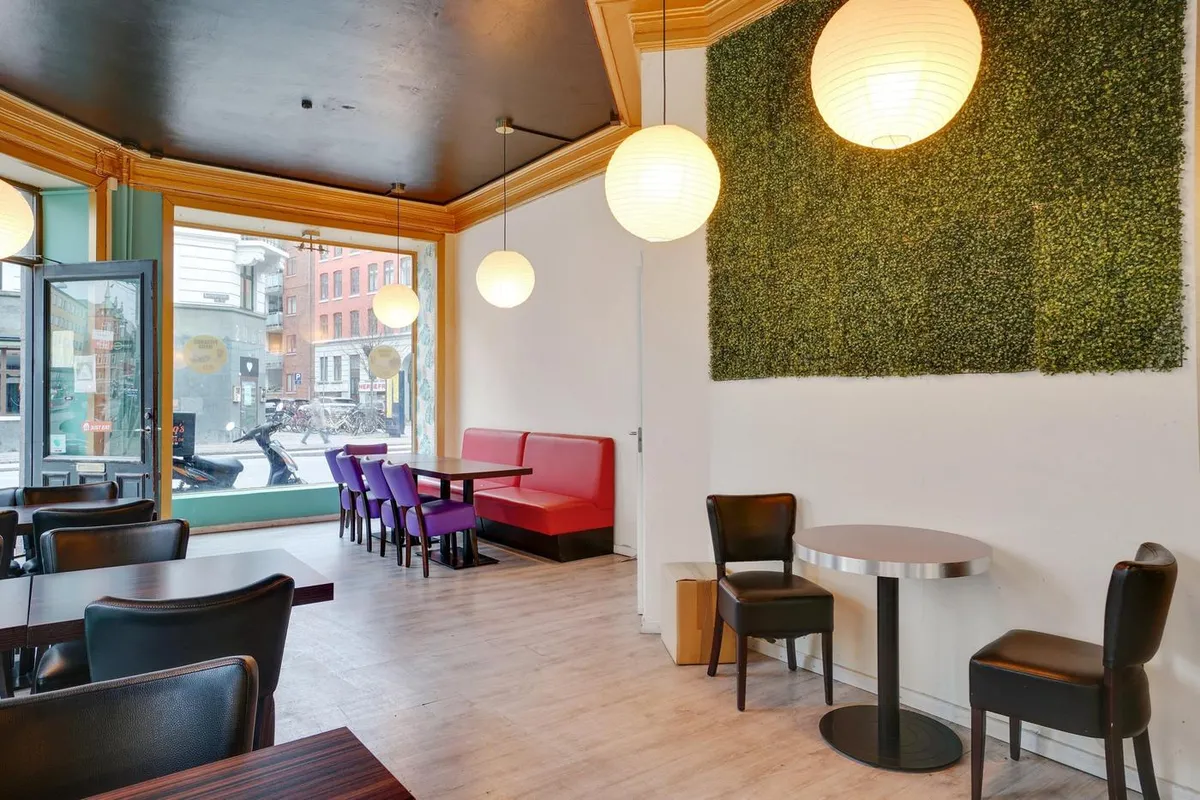 Moderne café med grønne vægbeklædning og runde lysekroner. Sorte stole og runde borde, en rødmalv hjørnesofa og et åbent