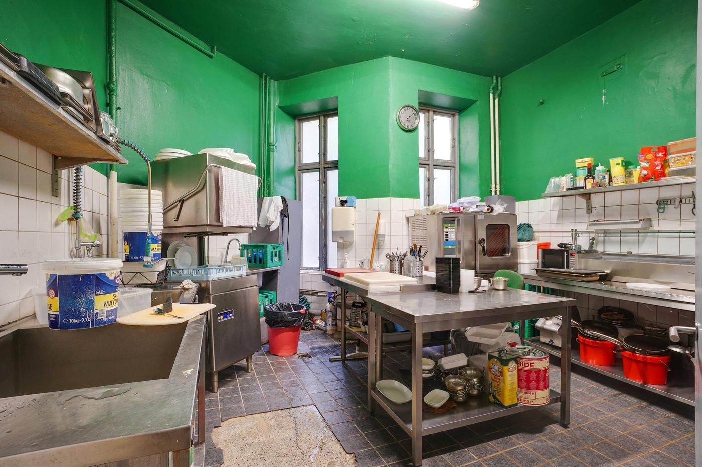 En køkkenrum med lyse grønne vægge, et køkkenbord med mad, en rulleserviet, flere gryder og gryder, og et vindue med ure