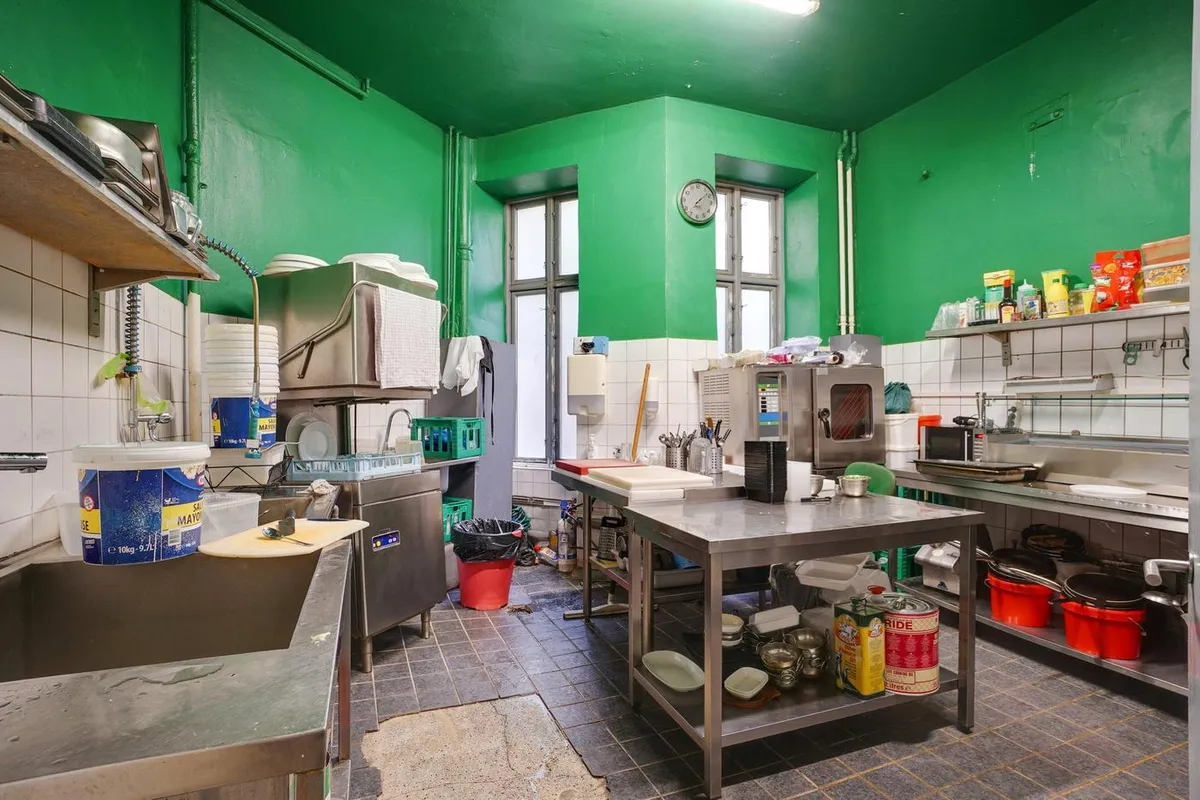 En køkkenrum med lyse grønne vægge, et køkkenbord med mad, en rulleserviet, flere gryder og gryder, og et vindue med ure