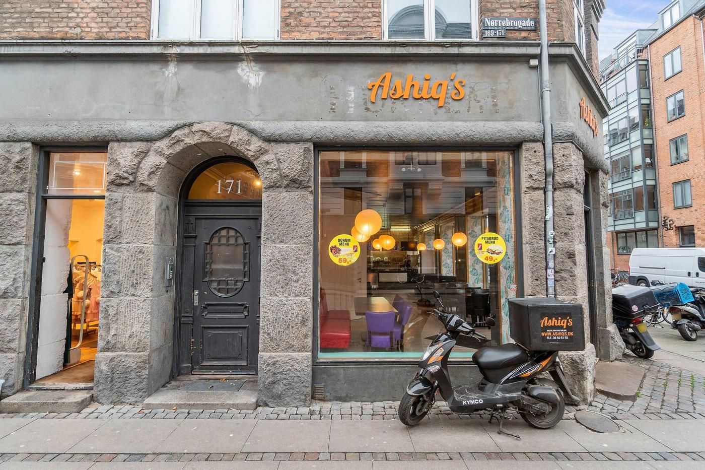 Et hus med butikker og en scooter parkeret udenfor på en stenbro. Butikkens dør er åben og mærket Ashiq's er synligt.