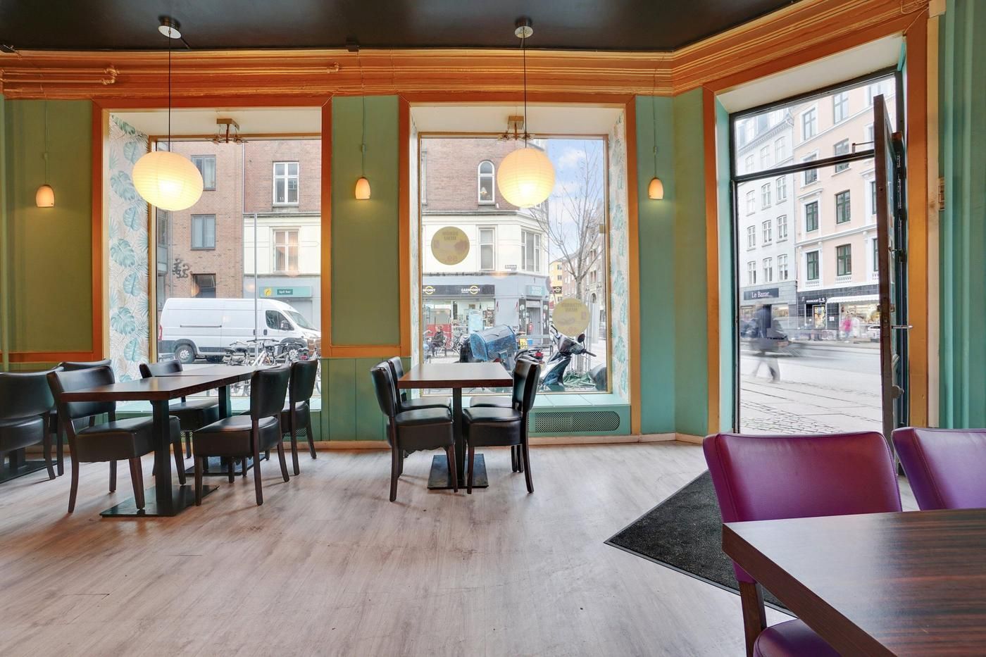 Inde i et café med borde og stole, lysekroner, grønne vægge og vinduer, uden folk og med byudsigt, samt en åben dør og et