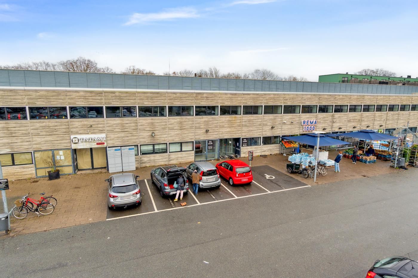 Rema 1000-butik med parkeringspladser, to røde biler, en person, der lægger indkøb, og to cykler udenfor