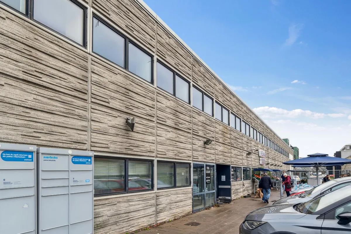 Butikslejemål med postkasser udenfor og biler parkeret udenfor, hvor to kunder går forbi en grøn paraply