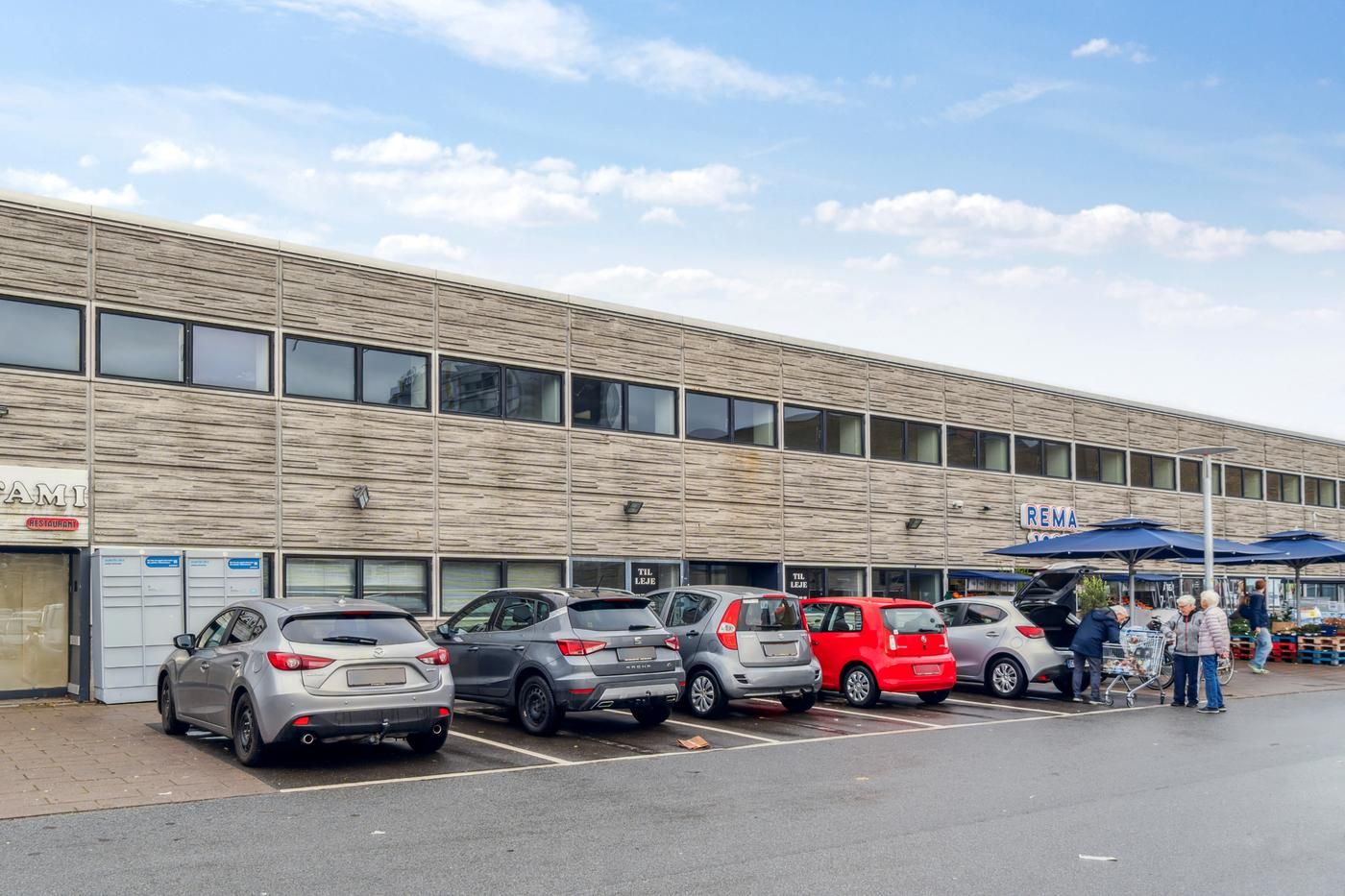 Flere biler parkeret foran Rema-supermarkedet og Rema 1000. Nogle mennesker går og skubber indkøbsvogne udenfor.