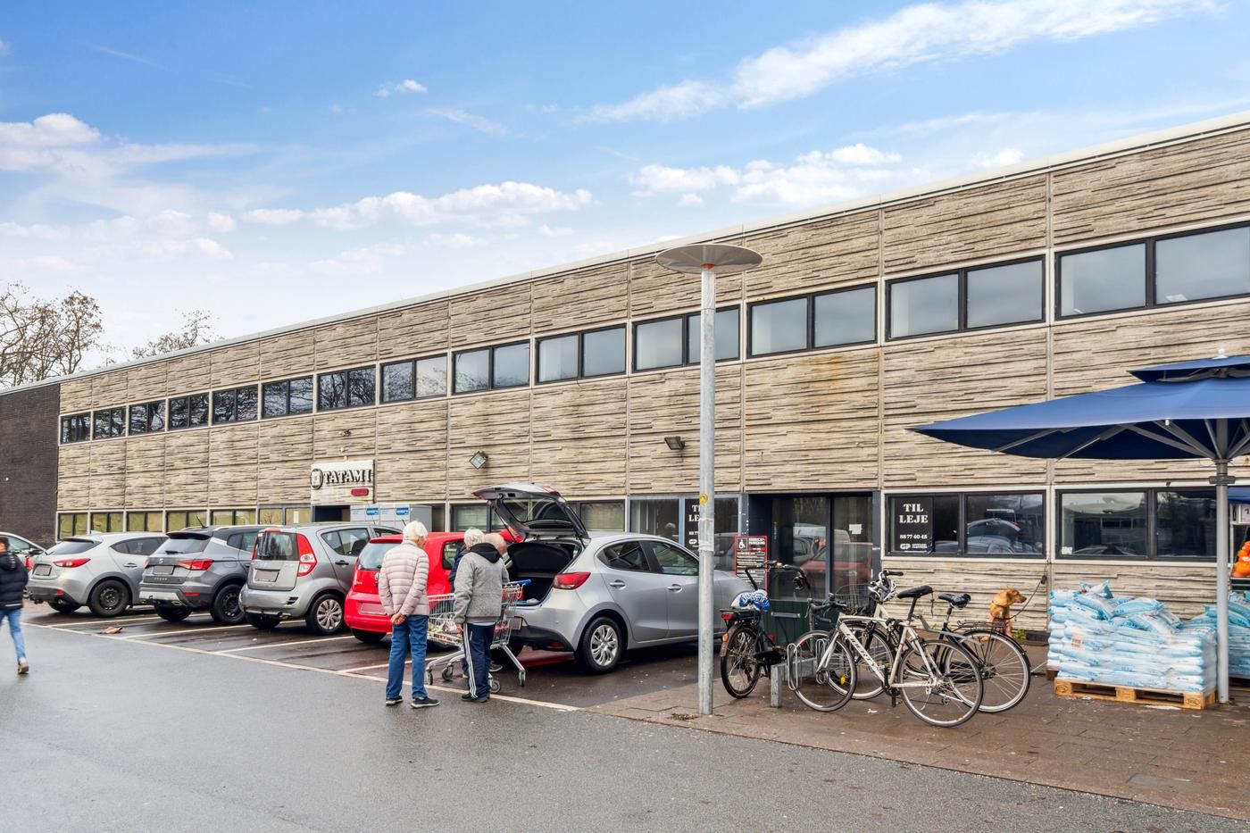 En parkeringsplads med parkerede biler og to personer, der laster indkøb i en bil med åben bagklappe og kørestole