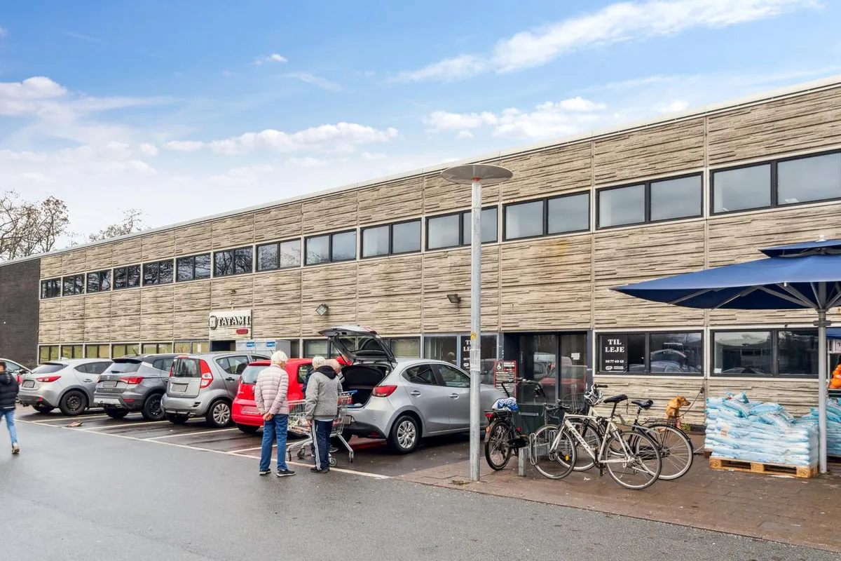 En parkeringsplads med parkerede biler og to personer, der laster indkøb i en bil med åben bagklappe og kørestole