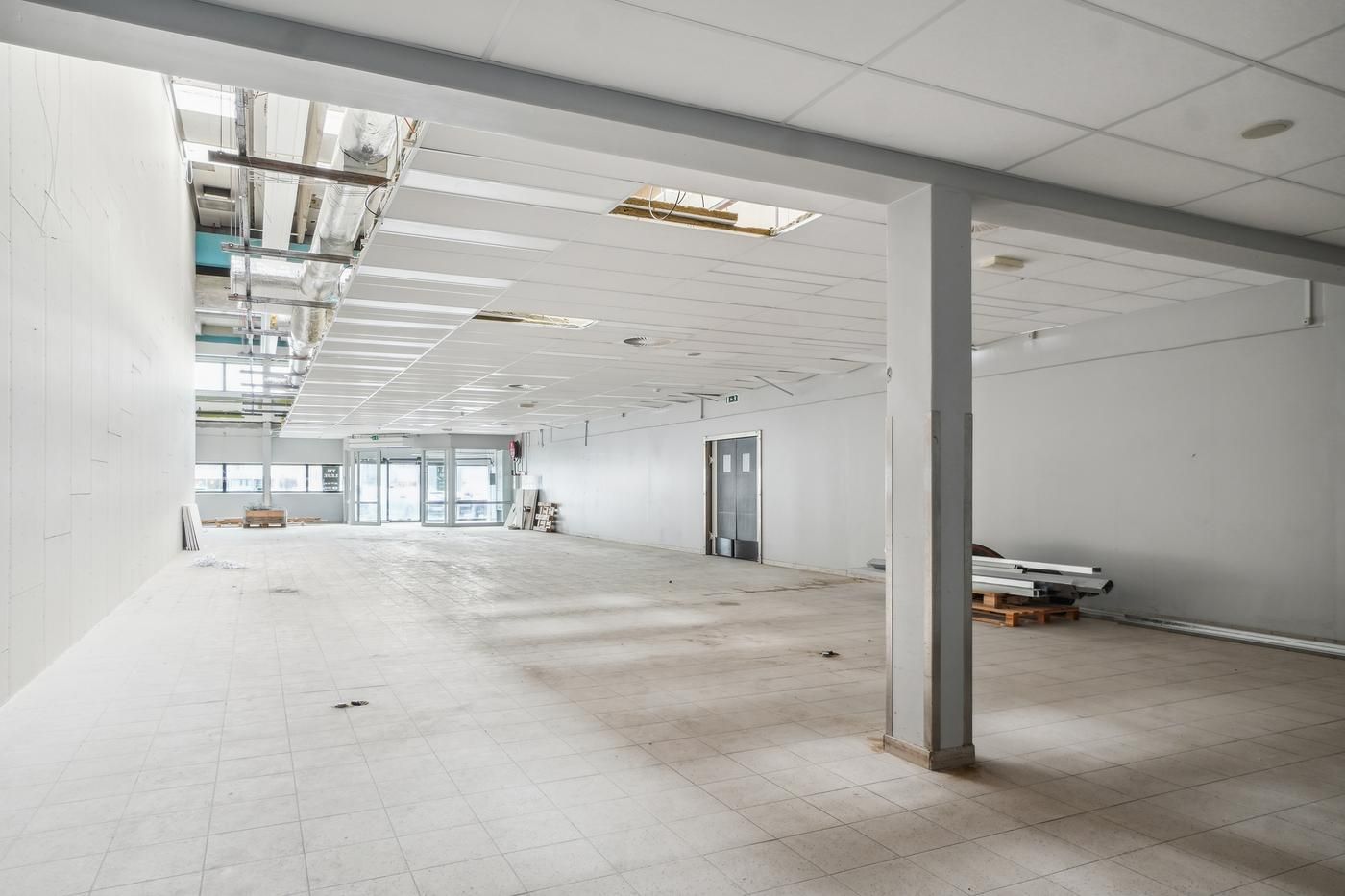 Ledig butikslokal med hvide vægge, højloft og glasdøre i et tomt lokale med solbelyst gulv og lagermateriale til højre