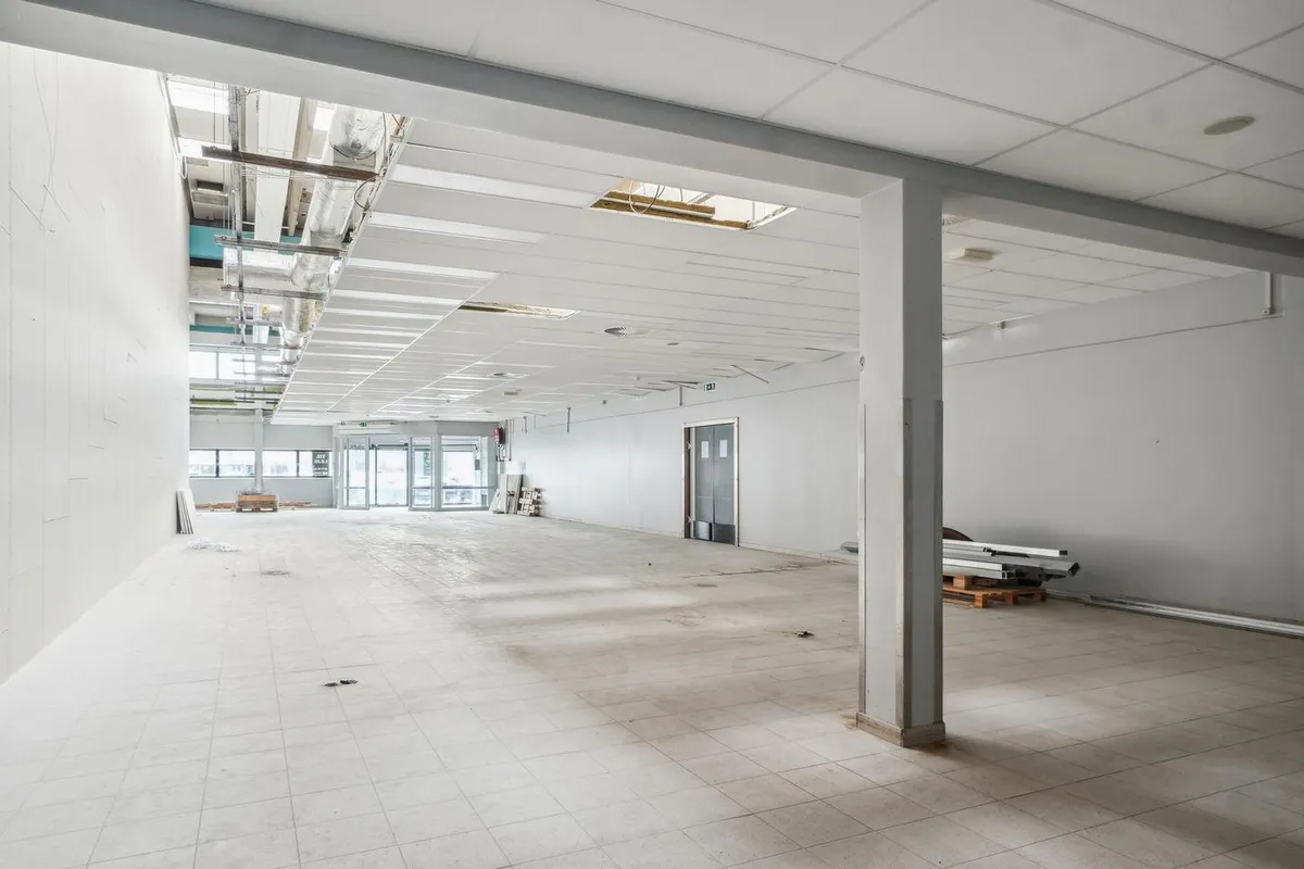 Ledig butikslokal med hvide vægge, højloft og glasdøre i et tomt lokale med solbelyst gulv og lagermateriale til højre