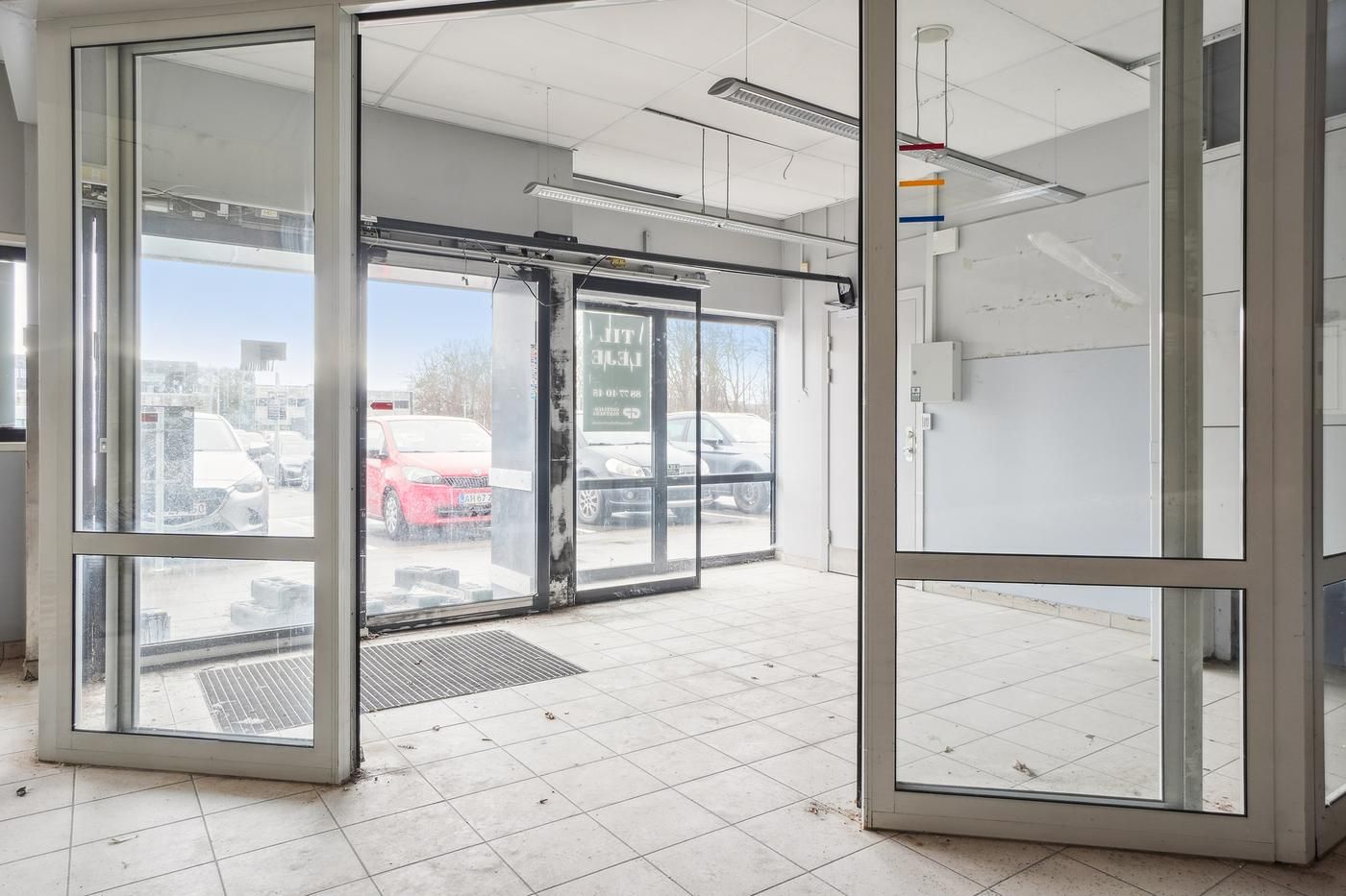 Lejemål med glasdør og gulvbelægning i forretningslokaler med åbne døre og biler udenfor i parkeringsområdet