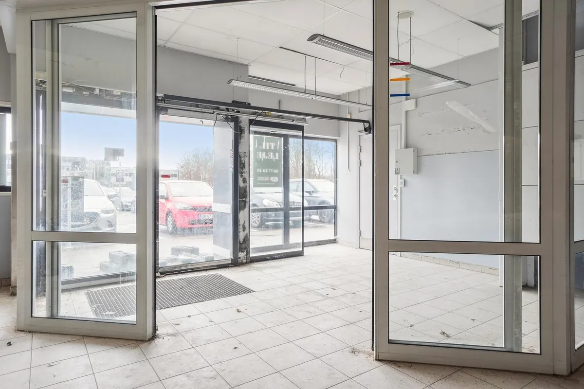 Lejemål med glasdør og gulvbelægning i forretningslokaler med åbne døre og biler udenfor i parkeringsområdet