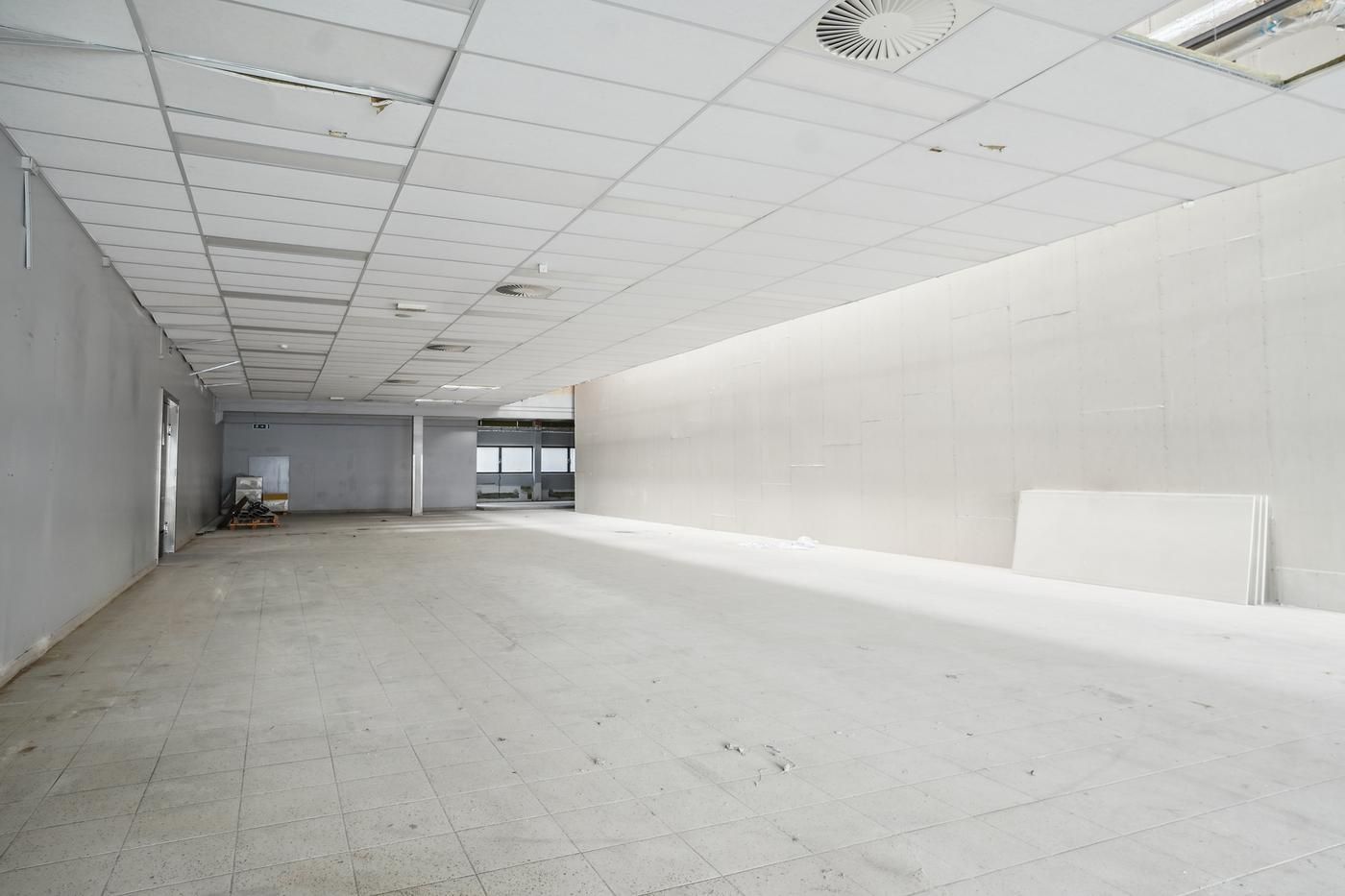 Rummelig tom butikslokal med hvide vægge og lofter, møbler og udstyr i baggrunden, ventiler og kasser til højre, gulv med