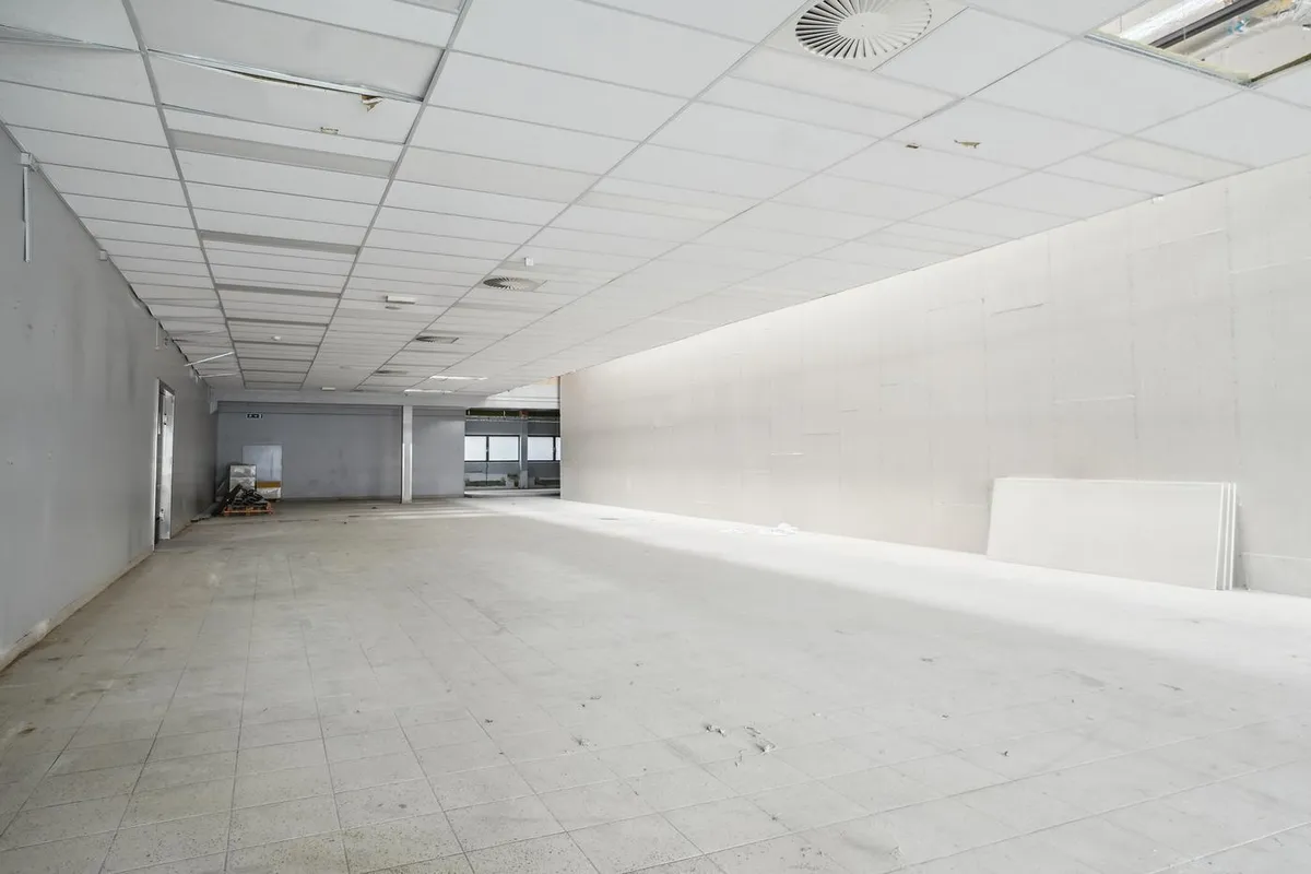 Rummelig tom butikslokal med hvide vægge og lofter, møbler og udstyr i baggrunden, ventiler og kasser til højre, gulv med